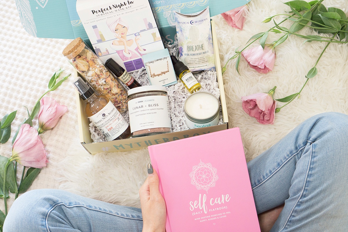 Seen from above, a woman holds a box that says My Therabox. It contains a pink self care book and several scented items.