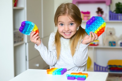 Smiling little  girl sitting at her white desk holding rainbow colored bubble pops in various shapes. 