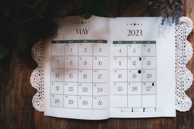 white calendar with black writing open and laid on a white crocheted placement on a wood table. shows calendar of may 