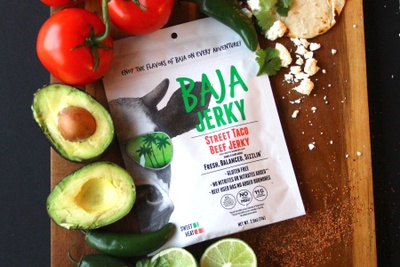 Baja Street Taco Jerky.  Bag is lying on a wooden cutting board with an open avocado, tomatoes, and lime slices surrounding it. 