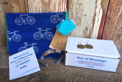 India Box of Blessings filled with Gold Arabesque Earrings, a silver cuff, and a Blue handbag with bicycles on it. 