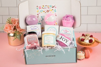 Treat Yourself Bath Bevy Box with bath bombs, whipped soap scrub, bubble rubble with flowers and pastries in the background
