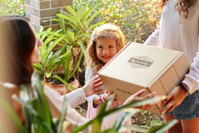 A woman handing a subscription box to another woman while a little girl looks on.
