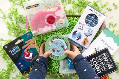 A young child's hands hold a round tin that says Regulation Putty over a subscription box with a spin cube and bath scrub.