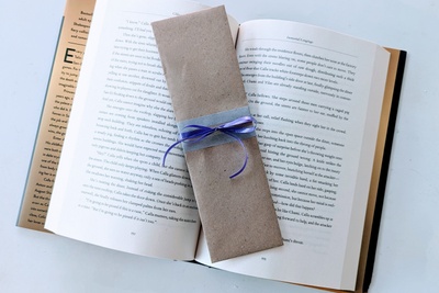 A Fantasy bookmark wrapped in brown craft paper and decorated with purple ribbon rests on an open hardcover book with golden edges. 