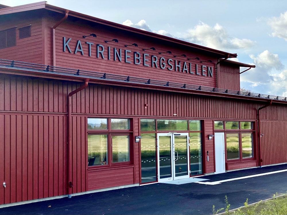 Entrén till en stor byggnad med röd träpanel, på fasaden ovanför ingången står texten "Katrinebergshallen". 
