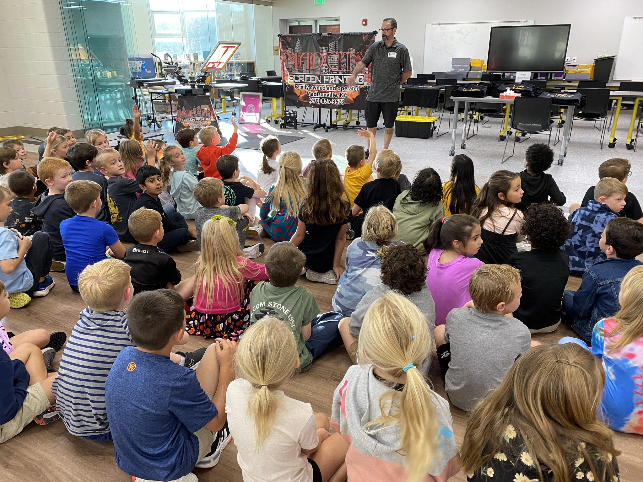 1 person stands at front of room, with more than 20 kids sitting on floor