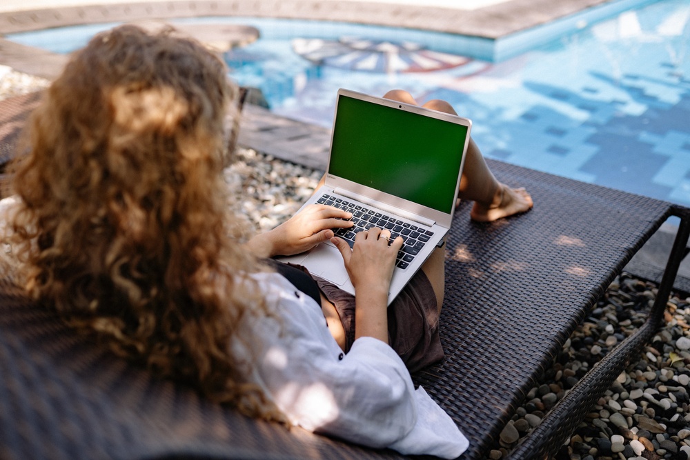 A red haired girl sitting in the shade by the pool, writing on her laptop.
