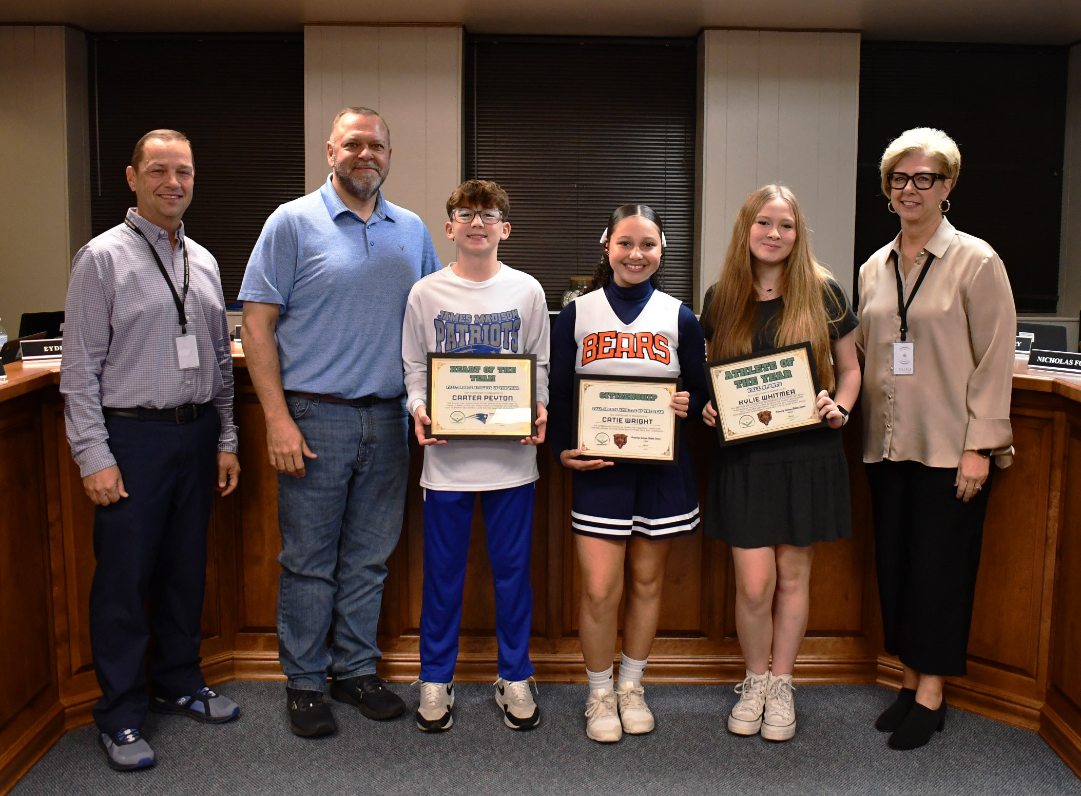 6 people stand in front of wooden furniture, with 3 holding framed certificates.