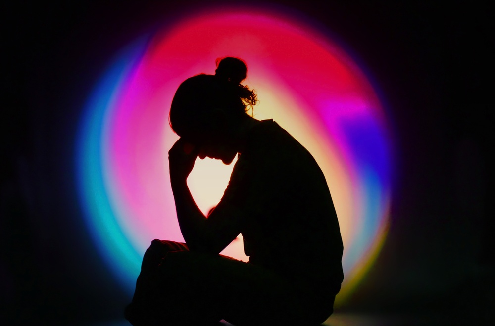 En person sitter som en siluett mot en svart bakgrund där en aura av ljus i regnbågens olika färger ligger som en cirkel i bakgrunden.