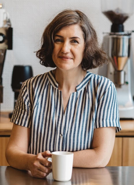 Headshot of Suzie Hoban, Head of Impact and Partnerships, Market Lane Coffee. Suzie is smiling at the camera in front of a coffee machine. She has a coffee mug in her hand.