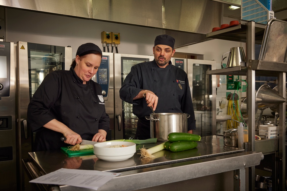 Bild på två kockar som arbetar i ett kök. På arbetsbänken i rostfritt stål ligger flera olika grönsaker, bland annat purjolök och squash.