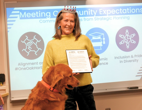 A woman wearing a crown holds a certificate in front of a whiteboard with community expectations listed.