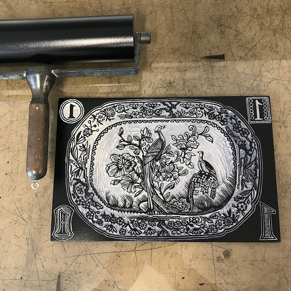 An inked up block with an intricate tree design with two pheasants and a floral border, positioned next to a black brayer.