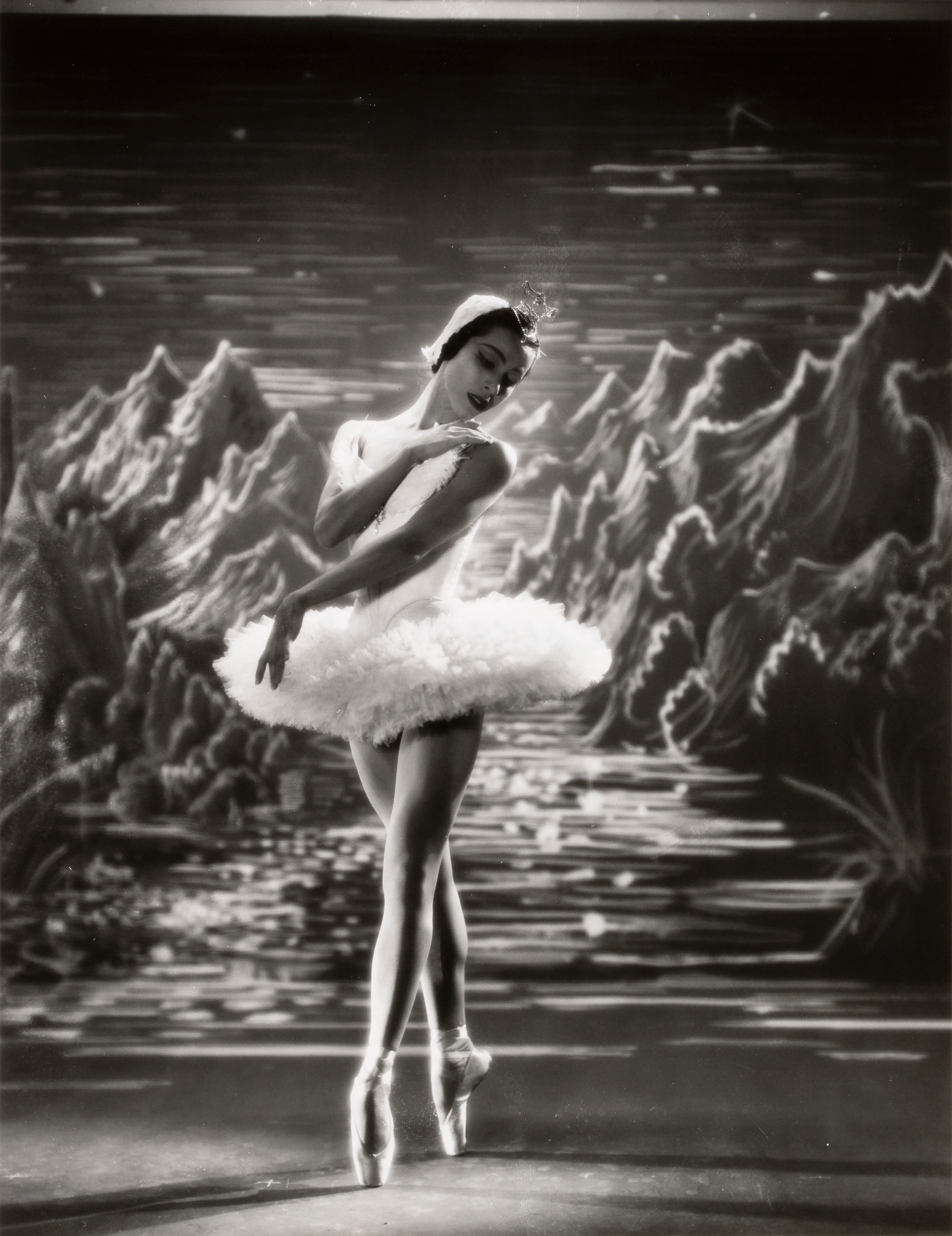 A black-and-white photograph of a ballet dancer in a tutu performing in front of an illustrated backdrop depicting a mountainous landscape.