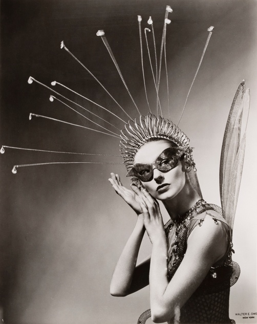 A black-and-white photograph portrait of a woman facing the camera wearing wings, a large headdress with protruding sticks and a mask around her eyes.