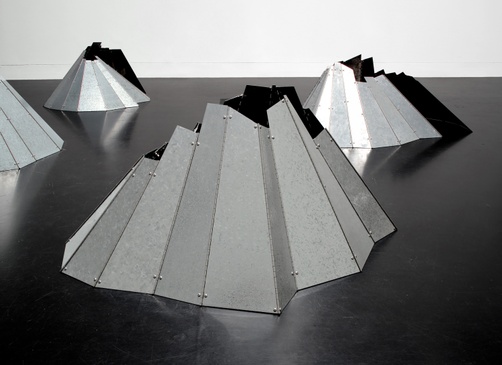 Five metal, silver, mountainous sculptures sit on a dark floor. 