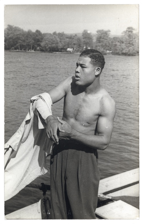 A black and white photograph of a young, black man taking off his shirt in front of a lake.