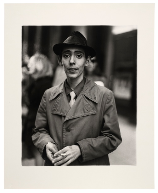 In a black and white portrait photograph, a young man wearing a trench coat and fedora hat stands holding a cigarette with an out-of-focus setting behind him.