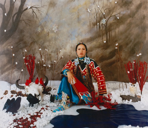 A young, tanned, Native woman sits in a fake snowy landscape wearing a bright red and blue dress with her hair in two braids.