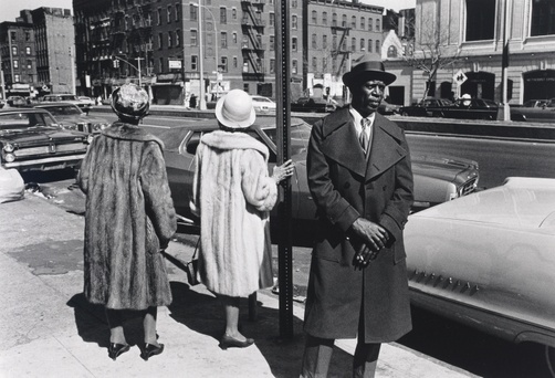 A black and white photograph of one black man and two black women standing on a street corner in a city.