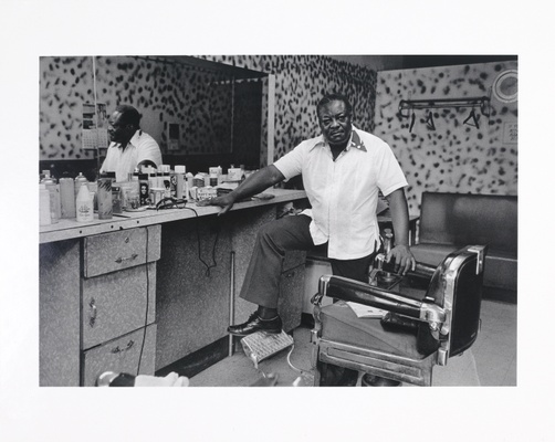 A black and white photo of a Black man with a white shirt standing in a barber shop.
