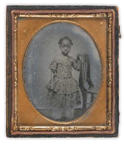 A black and white photograph of a young, black girl in a dress. The photograph is in a brown and bronze frame.