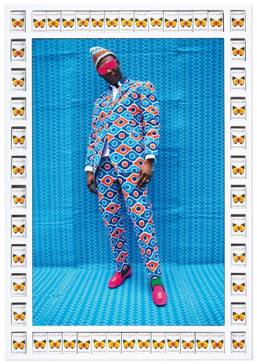 A photograph of a Black man in a blue and orange patterned suit with pink sunglasses and shoes standing in front of a blue patterned background in a frame lined with butterfly matchboxes.