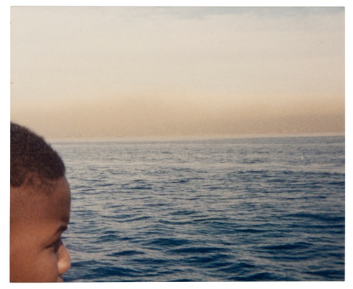 A dark-skinned boy, his head partially-cropped by the frame of the image, looks out on a body of water.