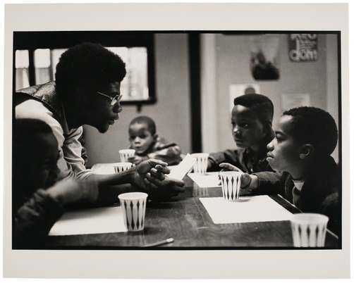 A Black man sits at a table and speaks to three young boys.