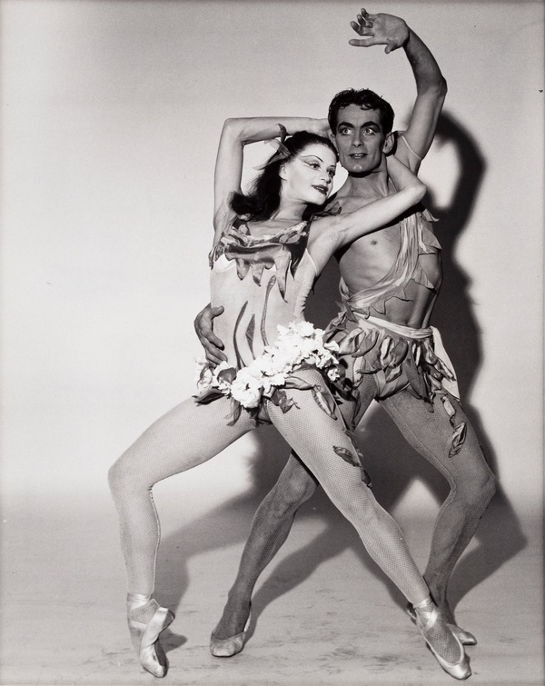 A black-and-white photograph of a man posing behind a lunging woman, both wearing small, elaborate costumes with flowers and fabric scraps.