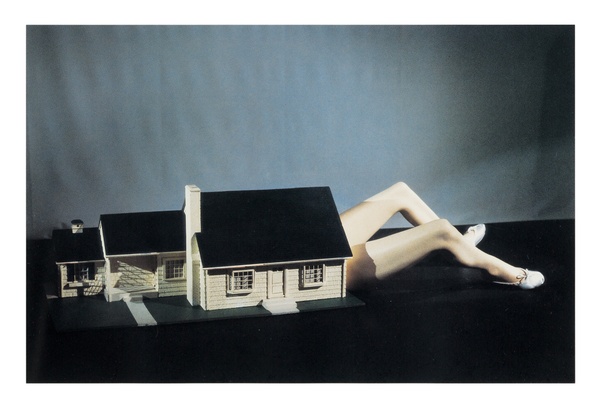 A three-part miniature house with a life-size pair of bare legs extending from one side. White slippers on the feet contrast against the black floor and gray background.