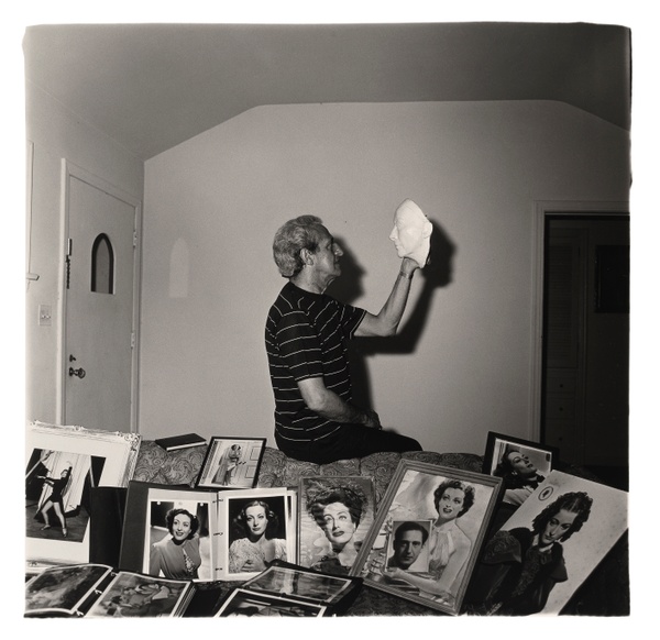 In a black and white photograph, a man sits perched on the back of a couch covered in pictures of Joan Crawford while holding and staring at a ceramic face.