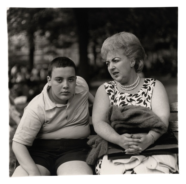 In a black and white photograph, a young boy intensely looks at us while sitting on a bench next to his mother who looks to the right.