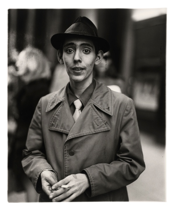 In a black and white portrait photograph, a young man wearing a trench coat and fedora hat stands holding a cigarette with an out-of-focus setting behind him.