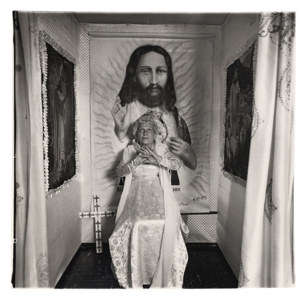 In a black and white photograph, a woman in a white dress looks up and crosses her hands in front of her chest in a small room featuring tapestries of Jesus.
