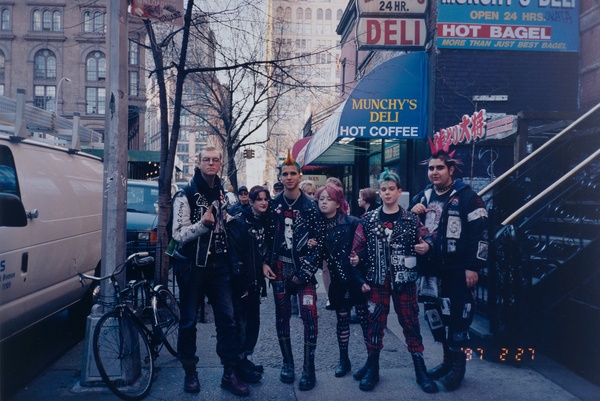 A group of six light-skinned people wearing black leather, spikes, piercings, and colorful hair stand side by side on a city sidewalk.