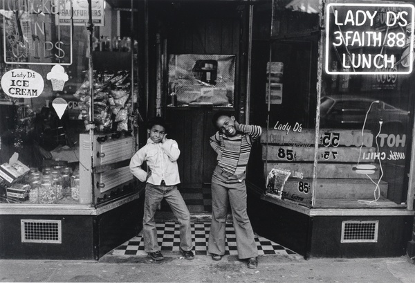 Two Black girls pose with their hands on their hips in front of an ice cream shop with an illuminated sign that reads “Lady D’s 3 Faith 88 Lunch.”