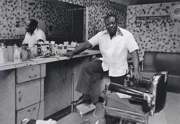 A black and white photo of a Black man with a white shirt standing in a barber shop.