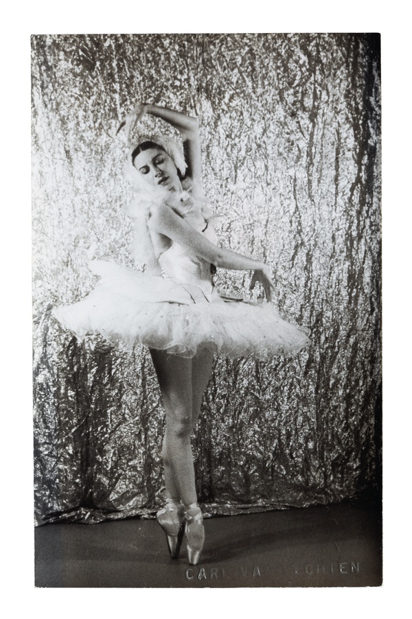A black and white photograph of a light-skinned, female ballet dancer in a white dress posing with one arm raised above her head.