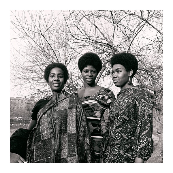 A black and white photograph of three smiling Black women in patterned clothing standing outside in front of a bare tree.