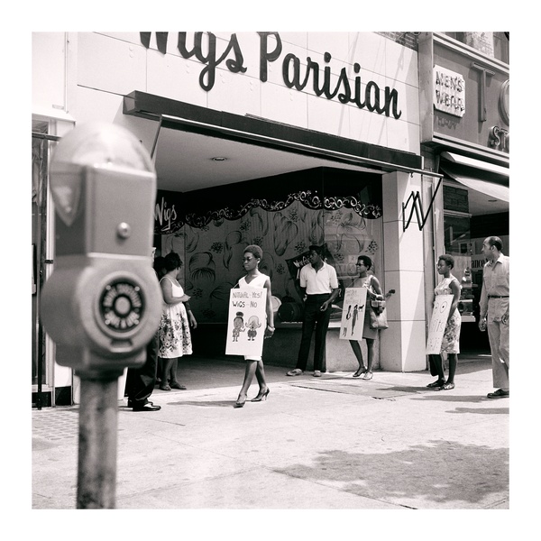A black and white photograph of three Black women with afros protesting with pro-natural hair signs outside a wig store while a few people watch from nearby on the sidewalk.