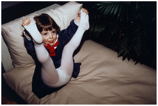 Sitting on a couch, a little girl playfully sticks her tongue out while pointing her legs up and grabbing her her toes, revealing white tights and underwear underneath her dress.