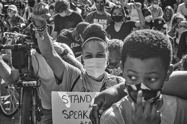 A black and white photograph of a crowd of protestors centered around a young, dark-skinned female wearing a mask, holding a sign and raising her right fist in the air.