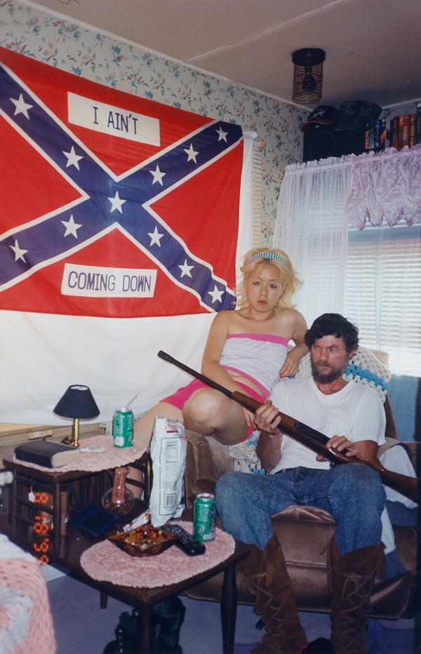 In a living room hangs a confederate flag that reads “I AIN’T COMING DOWN,” below which sits a Korean woman with dyed-blonde hair next to a white male holding a rifle. 