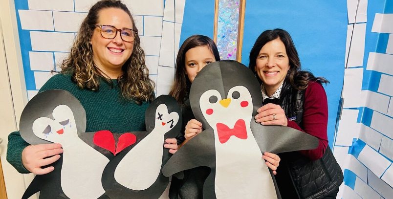 Two adults and a child pose with penguin cutouts, one with a broken heart, one with a sad face, and one with a bow tie.