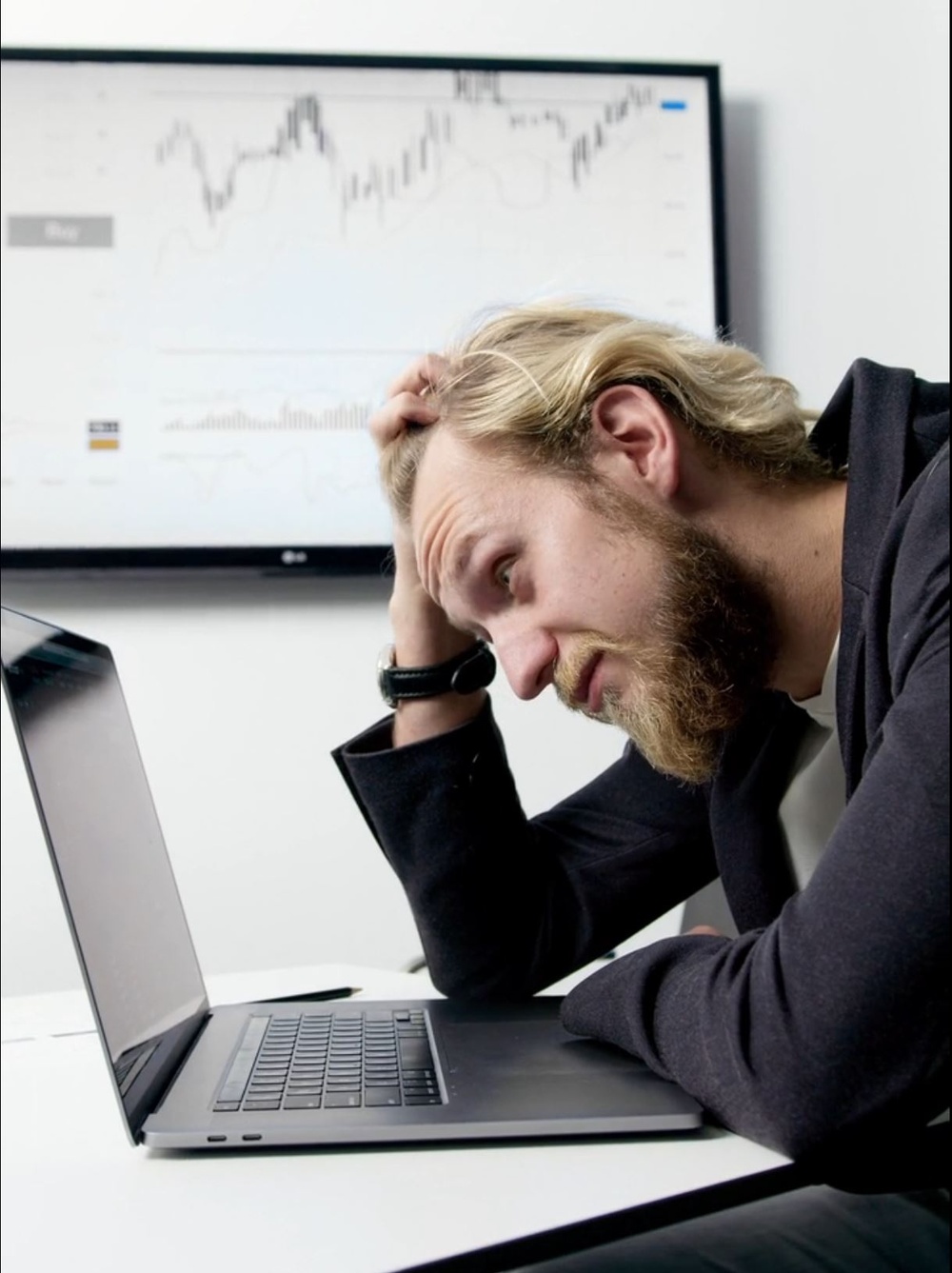 A blonde, bearded guy in a dark suite sits infront of his laptop pulling his hair and looking worried, in hte backgroudn there i a chart on the wall.