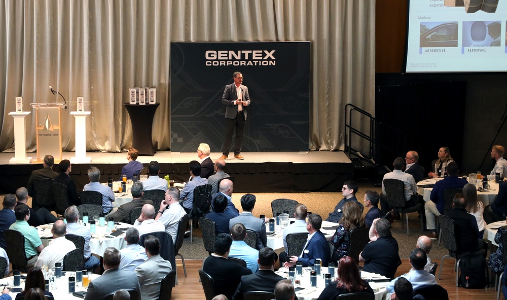 Man standing on stage with Gentex sign behind him. People sitting in chairs at round tables in front of him. 