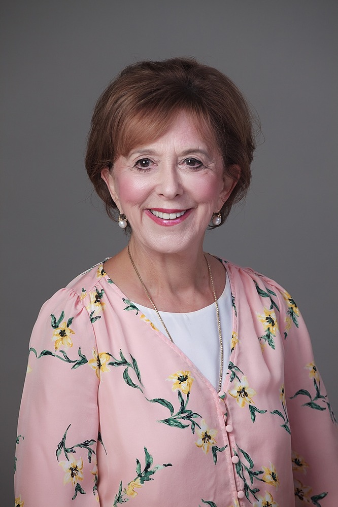 A person wearing a pink floral top is smiling at the camera in front of a grey background.