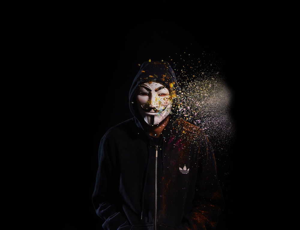 A black room and a human figure wearing a hoodie and an "anonymous" mask.